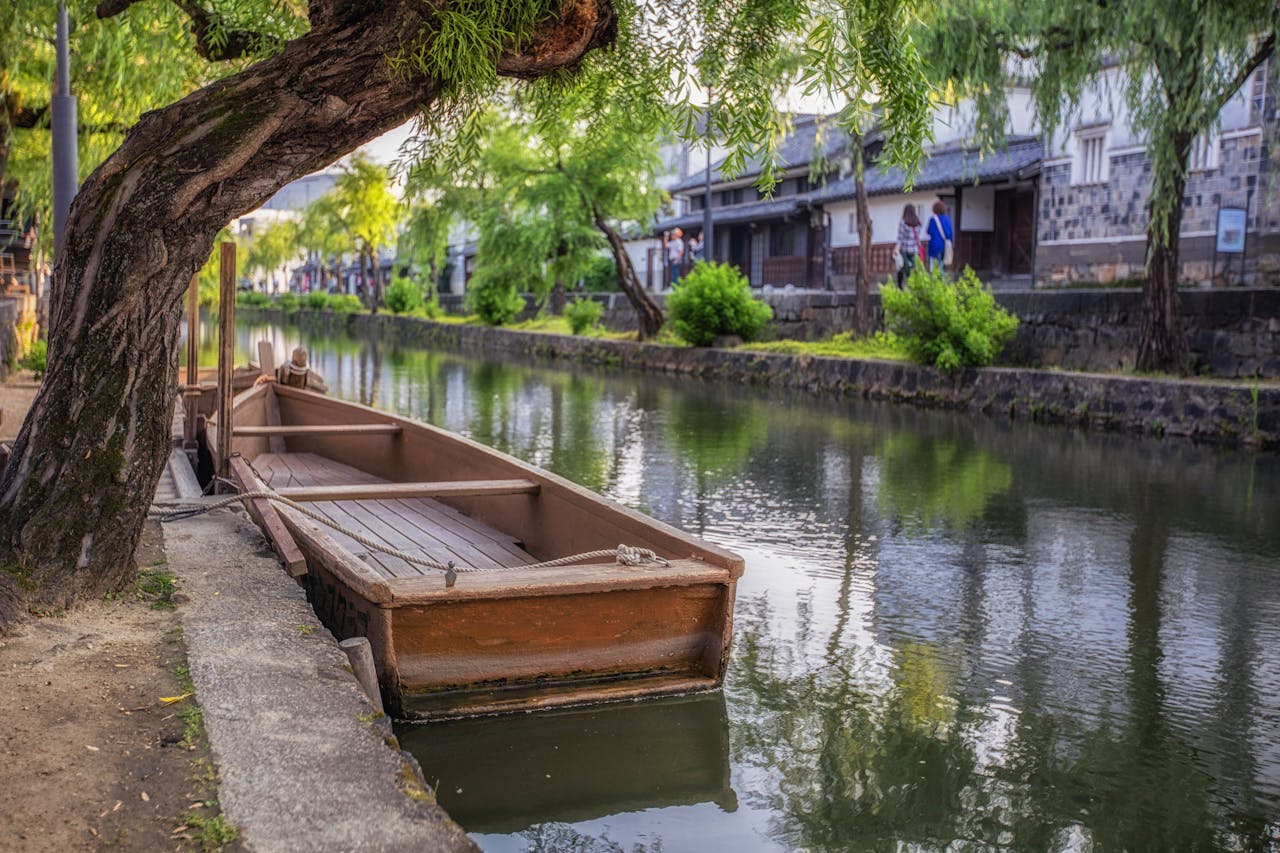 services-bg Scenic view of a traditional wooden boat docked along the canal in Kurashiki, Japan, surrounded by lush greenery.